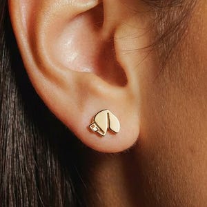 May include: Gold-colored stud earrings in the shape of a fortune cookie, one with a small, clear gem. The earrings are worn on an ear, with the focus on the jewelry. The earrings are a simple, elegant design.