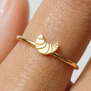 Könnte beinhalten: Ein dünner Goldring mit einem kleinen, halbmondförmigen Anhänger, der mit horizontalen Linien graviert ist. Der Anhänger befindet sich auf der Oberseite des Rings, der eine glatte, polierte Oberfläche aufweist. Der Ring wird an einem Finger getragen.