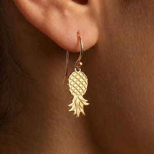 Può includere: Orecchini a forma di ananas color oro. Gli orecchini presentano un design dettagliato di ananas con squame strutturate e una corona di foglie. Gli orecchini sono appesi all'orecchio di una persona. Gioielli a tema tropicale.