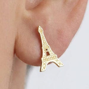 May include: A gold-toned stud earring shaped like the Eiffel Tower. The metal piece features a cutout design that mimics the structural lattice of the landmark, with a pointed top and a wide base, secured directly against the earlobe.