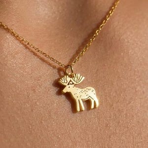 Puede incluir: Un collar dorado con un colgante con forma de alce. El colgante tiene cuernos detallados y un patrón punteado en su cuerpo. La delicada cadena también es dorada, creando un diseño simple y elegante. Joyería para uso diario.