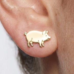 Könnte beinhalten: Ein goldfarbener Ohrstecker in Form eines Schweins im Profil. Das Schmuckstück hat eine glatte, polierte Oberfläche mit der einfachen Kontur eines Schweins, einschließlich eines kleinen geringelten Schwanzes, vier Beinen und einem Rüssel.