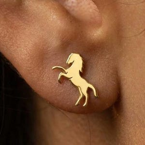 May include: A gold-colored stud earring in the shape of a rearing horse is shown on an ear. The earring is a simple, elegant design, with the horse's silhouette clearly defined. The earring is a small, delicate piece of jewelry.