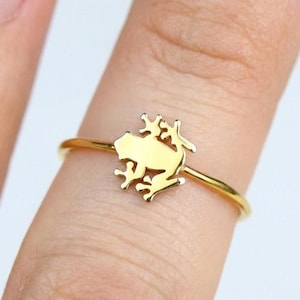 May include: A gold-toned ring featuring a small, flat, stylised frog silhouette centred on a thin, smooth metal band. The frog design is detailed with cutouts representing limbs and a head, creating a minimalist and polished metallic appearance.
