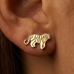 May include: Gold-colored tiger stud earrings. The earrings feature a detailed tiger design with stripes. The earrings are small and are worn on the earlobe. The earrings are a jewelry item.