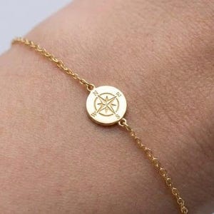 May include: A delicate gold-tone chain bracelet featuring a small, circular charm in the center. The charm is engraved with a detailed compass rose design, including the cardinal direction letters N, S, E, and W etched into the metal surface.