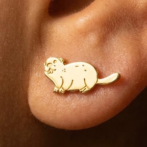 May include: A gold-colored stud earring in the shape of a groundhog is shown on an ear. The groundhog has a smiling face and a small tail. The earring is a small, detailed piece of jewelry.
