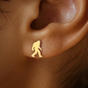 May include: A gold-colored stud earring in the shape of a Bigfoot silhouette. The earring is small and simple, with a smooth surface and a minimalist design. The earring is worn on the ear.