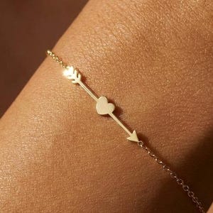 May include: A delicate gold chain bracelet featuring a central horizontal arrow motif integrated with a small heart shape. The metal has a polished finish, and the design consists of a thin link chain connected to the arrow and heart centrepiece.