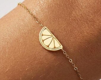 May include: A delicate gold chain bracelet featuring a small, flat, semi-circular charm shaped like a citrus slice. The charm has etched lines radiating from the center to the curved edge, creating a segmented appearance on the polished gold surface.