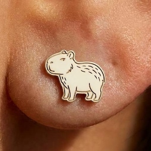 May include: A gold-colored stud earring featuring a cream-colored capybara design. The capybara is outlined in black and has detailed markings. The earring is worn on an ear.