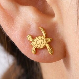 May include: A gold-toned stud earring shaped like a sea turtle with a detailed shell pattern. The earring is positioned on the earlobe, featuring a polished metallic finish and a small, flat design that sits flush against the skin of the ear.