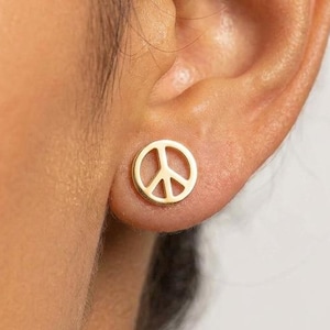 May include: A small, circular gold stud earring featuring a cut-out peace sign symbol. The earring is worn in the earlobe, displaying a polished metallic finish and a simple, clean design that sits flush against the skin of the ear.