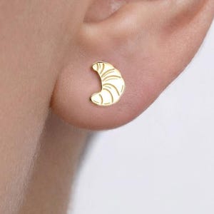 May include: A small gold stud earring shaped like a croissant. The earring features a polished metallic finish with engraved lines that mimic the layered texture of the pastry. It is positioned on the earlobe, showcasing a simple and compact design.