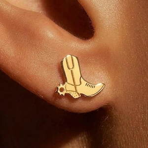 May include: A gold stud earring shaped like a cowboy boot with a small spur detail on the heel. The earring features a polished metallic finish with engraved lines defining the boot shaft, toe, and heel, securely positioned in an earlobe.