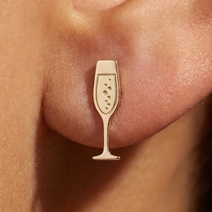 May include: A gold-colored champagne flute stud earring. The earring is a small, detailed representation of a champagne flute with bubbles. The earring is worn on the ear. The overall theme is jewelry.