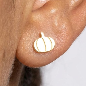 May include: A small, gold-toned stud earring shaped like a pumpkin. The earring features a smooth, polished surface with vertical engraved lines that define the segments of the pumpkin shape, creating a simple and distinct silhouette on the earlobe.