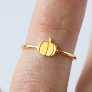 Può includere: Un sottile anello color oro caratterizzato da un piccolo ciondolo a forma di zucca centrato. Il design della zucca presenta linee verticali incise per definirne la forma, e la fascia è un filo circolare semplice, liscio e lucido.