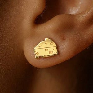 May include: A small gold stud earring shaped like a wedge of Swiss cheese. The earring features a flat, triangular design with several circular cutouts representing holes, finished in a polished metallic gold tone that catches the light.
