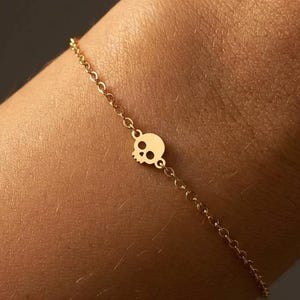 May include: A delicate gold-colored bracelet featuring a small skull charm. The charm is centered on a fine chain, with an adjustable length. The bracelet is shown on a wrist against a dark background.