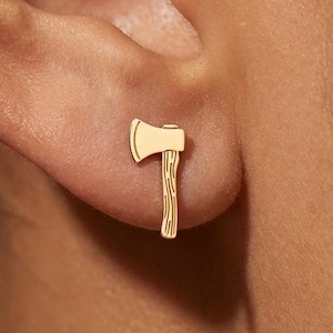 May include: A gold-colored axe stud earring. The earring is a small, detailed representation of an axe, with a blade and a wooden handle. The earring is worn on the ear.