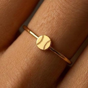 May include: A delicate gold ring featuring a small, circular baseball design. The ring's band is thin and simple, and the baseball detail is etched with stitching lines. The ring is shown on a finger, highlighting its elegant and sporty design.