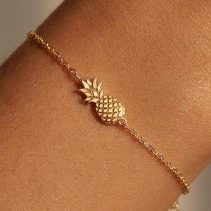 Puede incluir: Un brazalete dorado con un dije de piña. El brazalete de cadena fina tiene una longitud ajustable. El dije de piña está detallado con una superficie texturizada. El brazalete se usa en la muñeca.
