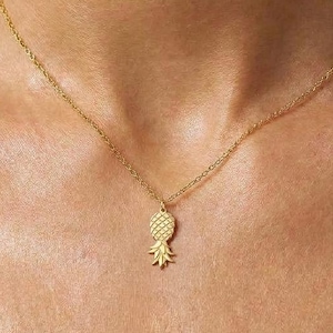 Puede incluir: Un collar de tono dorado que presenta una cadena delicada y un pequeño colgante de piña texturizado. El colgante muestra un patrón de cuadrícula en el cuerpo y hojas puntiagudas en la parte superior, suspendido centralmente de la cadena de metal pulido.