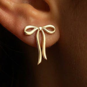 Puede incluir: Primer plano de un pendiente de botón dorado con forma de lazo. El pendiente tiene un diseño sencillo y elegante con un lazo y dos colas colgantes. El pendiente se lleva en una oreja.