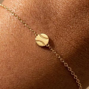 May include: A delicate gold bracelet featuring a small baseball charm. The charm is round with stitched detailing, and the bracelet has a heart-shaped tag. The bracelet is worn on a wrist.