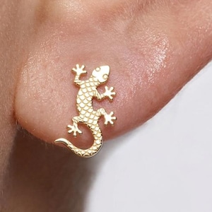 Puede incluir: Un pendiente de botón dorado con forma de pequeño lagarto con una superficie texturizada y escamosa. El lagarto se muestra en una posición curva con la cola hacia adentro, presentando cuatro patas bien definidas y una cabeza detallada.