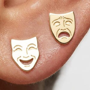 May include: A pair of gold stud earrings featuring the comedy and tragedy theater masks. One earring depicts a smiling face with upturned eyes and a wide mouth, while the other earring shows a frowning face with downturned eyes and a sad mouth.