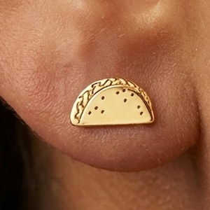 May include: A small gold-toned stud earring shaped like a taco. The earring features a curved top edge with a textured pattern representing the shell and a smooth, flat bottom section with small engraved dots across the surface of the taco.
