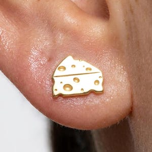 May include: A gold-toned stud earring shaped like a wedge of Swiss cheese, featuring a smooth surface with several circular cutouts of varying sizes. The earring is positioned on an earlobe, showcasing its flat, triangular design and metallic finish.