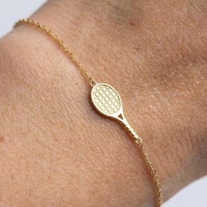 May include: A delicate gold chain bracelet featuring a small, centered tennis racket charm. The charm has a detailed grid pattern on the head of the racket and a slender handle, connected to a thin, polished gold link chain that encircles the wrist.