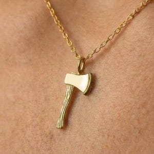 Pode incluir: Colar com pingente de machado dourado. O pingente apresenta um design detalhado de machado com uma lâmina creme e um cabo texturizado. O colar é uma corrente delicada, perfeita para o uso diário.