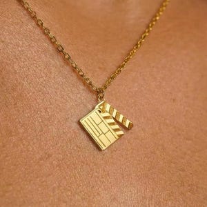 Puede incluir: Un collar de tono dorado con un pequeño colgante plano en forma de claqueta de cine. El colgante está suspendido de una cadena fina y delicada de tono dorado. El dije de claqueta tiene líneas grabadas que representan el tablero y el brazo.