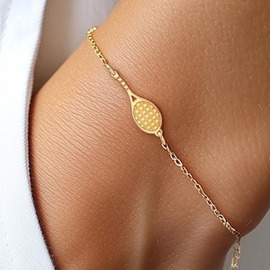 May include: Gold-colored tennis racket charm bracelet. The delicate chain bracelet features a small tennis racket pendant and a heart-shaped charm at the adjustable clasp. The bracelet is worn on a wrist.
