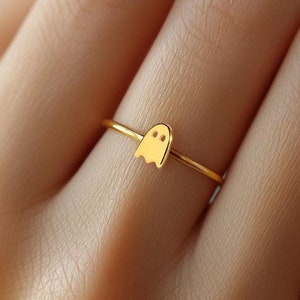 Könnte beinhalten: Ein goldener Ring mit einem kleinen Geister-Anhänger. Der Ring wird an einem Finger getragen.