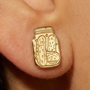 Pode incluir: Um brinco de pino dourado em forma de frasco retangular contendo dois picles. O design apresenta linhas gravadas que detalham a tampa do frasco, a textura dos picles no interior e o contorno do recipiente em acabamento de metal polido.