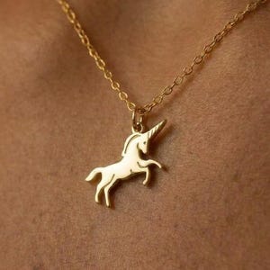 May include: A gold-toned necklace featuring a delicate chain and a pendant shaped like a prancing unicorn. The unicorn pendant is rendered in a smooth, polished gold-colored metal with defined outlines for the mane, tail, and horn, suspended from a link chain.