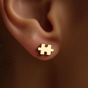May include: A gold-colored stud earring in the shape of a puzzle piece is shown on an ear. The earring is small and has a smooth surface. The background is blurred, focusing attention on the jewelry.