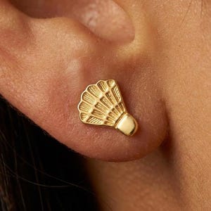 May include: A gold stud earring shaped like a badminton shuttlecock. The earring features a textured, fan-like top section with vertical ridges and a smooth, rounded base. The piece is polished and metallic, designed to be worn directly on the earlobe.