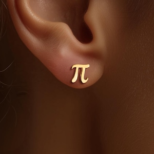May include: A close-up of a gold-colored stud earring shaped like the Greek letter pi, worn on an ear. The earring is small and simple, with a polished finish. The background is blurred, focusing attention on the jewelry.