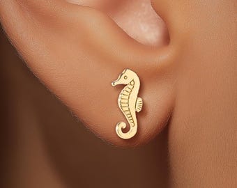 May include: A small, gold-toned stud earring shaped like a seahorse. The earring features a detailed, textured design with a curved tail and a defined head, securely positioned on the earlobe. The metal has a polished, reflective finish.