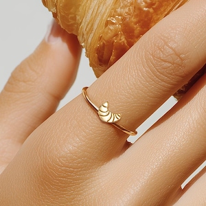 May include: A golden ring with a croissant design is worn on a finger, next to a fresh croissant. The ring is delicate and simple, with the croissant shape as the focal point. The croissant is golden brown and flaky.