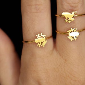 May include: Three gold-toned rings featuring a small, flat frog silhouette design. One ring is worn on the ring finger, while two rings are stacked on the middle finger. The rings have a thin, polished band and a smooth, reflective metallic finish.