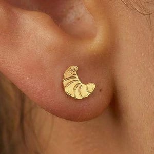 Puede incluir: Un pendiente de botón dorado con forma de croissant se muestra en una oreja. El pendiente tiene líneas detalladas para representar la textura del bollo. Una pequeña y delicada pieza de joyería.