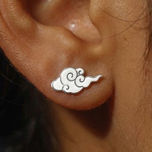 Puede incluir: Primer plano de un pendiente de botón plateado con forma de nube en una oreja. El pendiente tiene un diseño detallado y arremolinado dentro de la forma de la nube. Se ve la piel alrededor de la oreja.