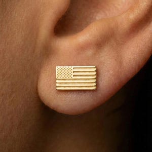 May include: A gold-colored stud earring shaped like the American flag, featuring horizontal stripes and a field of stars. The earring is rectangular and worn on an ear.